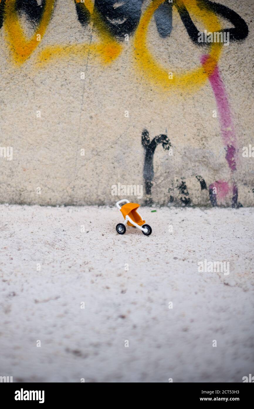 a closeup of a miniature plastic orange and white toy Playmobile pram on a concrete ledge by a wall with sprayed tags and graffiti in an urban environ Stock Photo