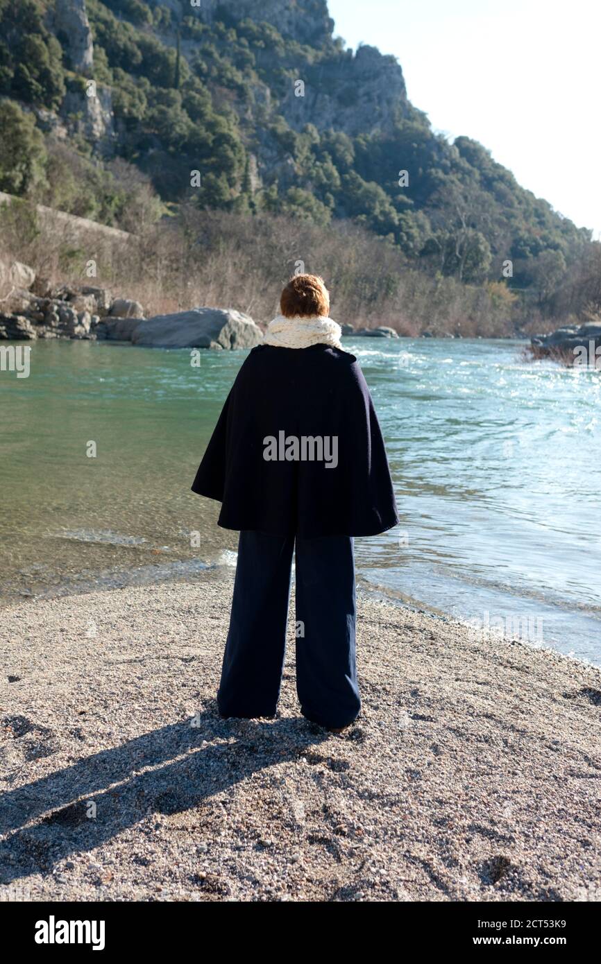 rear view of woman with short orange hair and dressed in stylish winter outfit stands on Herault river's pebble bank looking across the flowing river Stock Photo