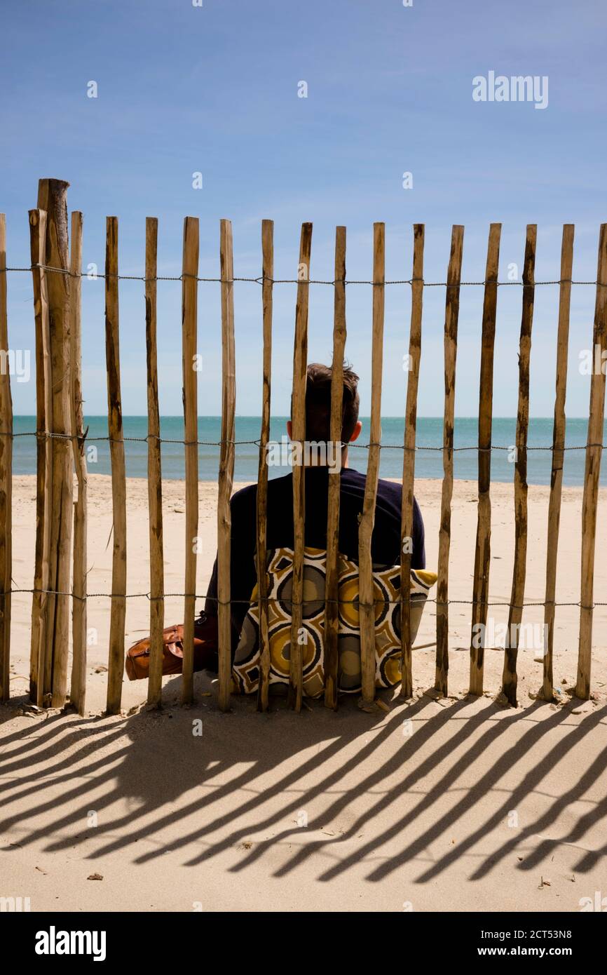 rear view of young man sitting on beach leaning against a vintage cushion and wooden fence and looking out across the mediterranean sea on a sunny day Stock Photo