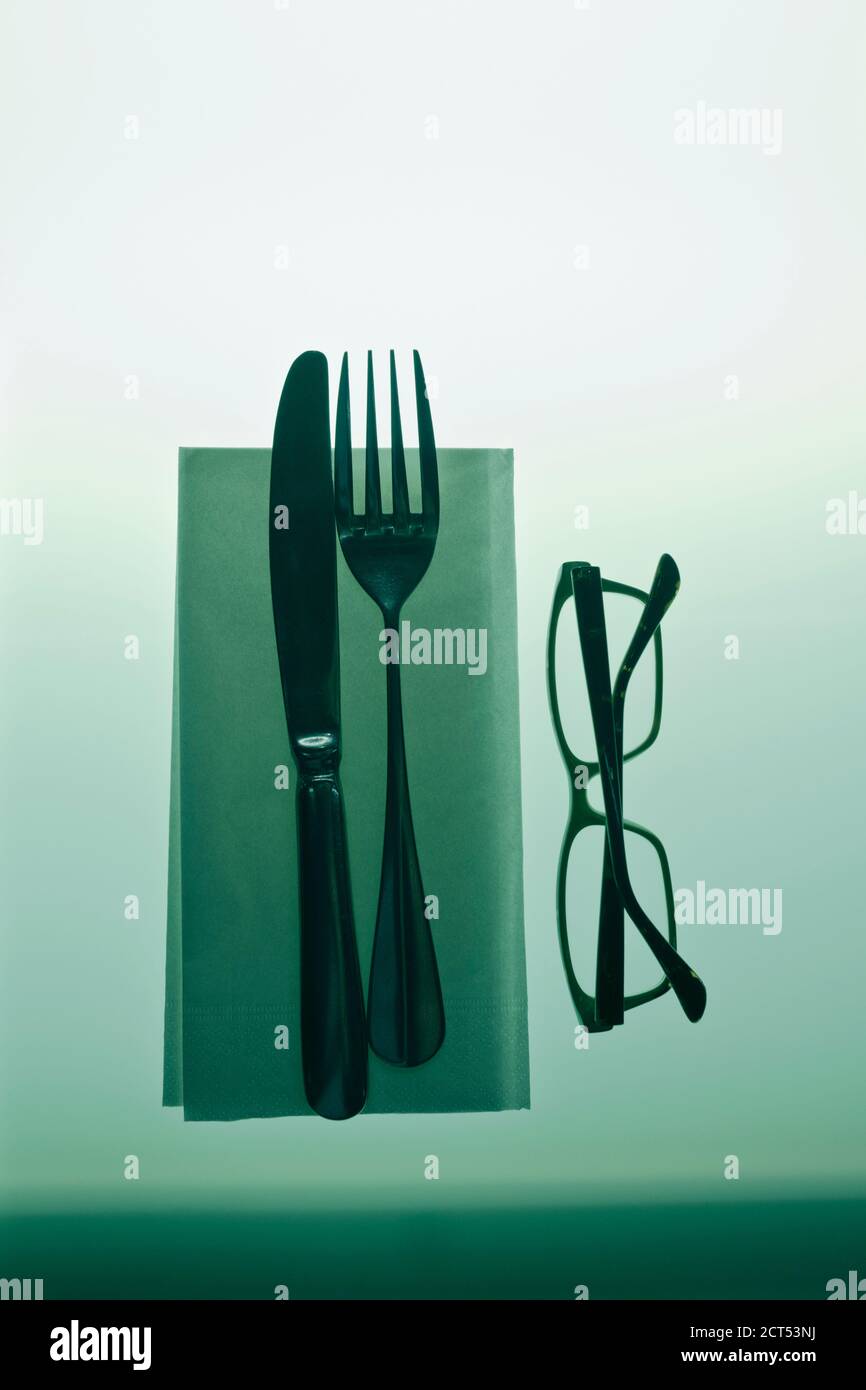 photo of knife and fork on a paper knapkin next to a pair of glasses on a backlight glass table Stock Photo