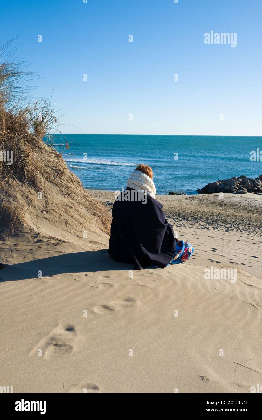rear view of woman dressed in winter outfit sitting in sand dunes on a beach looking out across the mediterranean sea on a sunny day Stock Photo