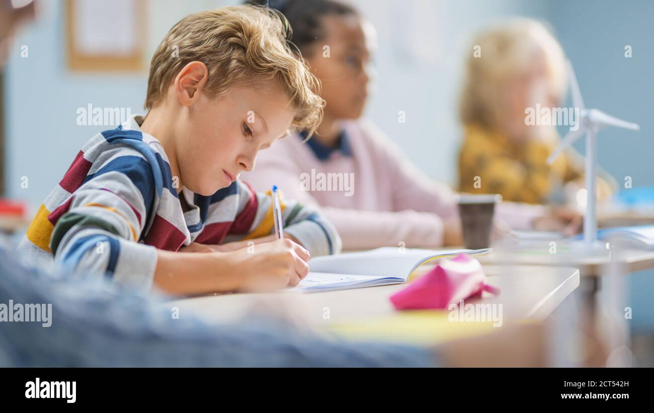 In Elementary School Classroom Brilliant Caucasian Boy Writes in Exercise Notebook, Taking Test and Writing Exam. Junior Classroom with Group of Stock Photo