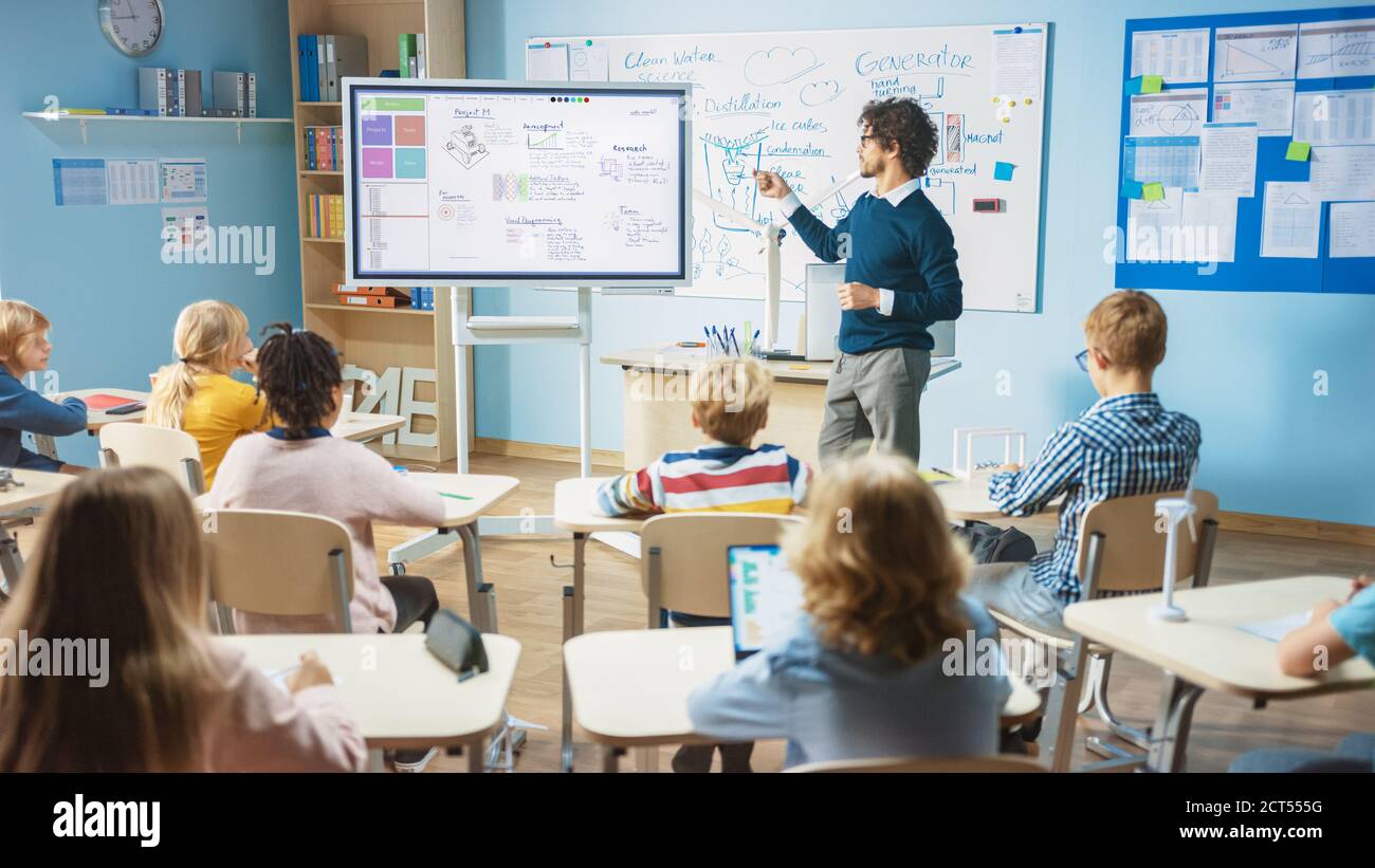 Elementary School Science Teacher Uses Interactive Digital Whiteboard to Show Classroom Full of Children how Software Programming works for Robotics Stock Photo