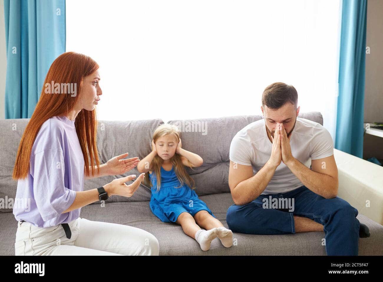 sad, desperate little girl during parents quarrel, she clog the ears sitting on sofa at home, angry parents fighting. worried upset small daughter hur Stock Photo