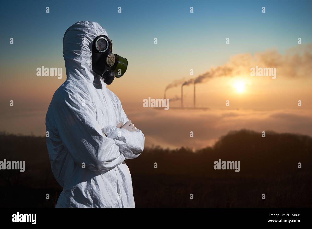 Side view of male environmentalist with crossed arms looking aside while thermal chimneys producing smoke with toxic gases into atmosphere. Ecologist wearing white radiation suit and gas mask. Stock Photo