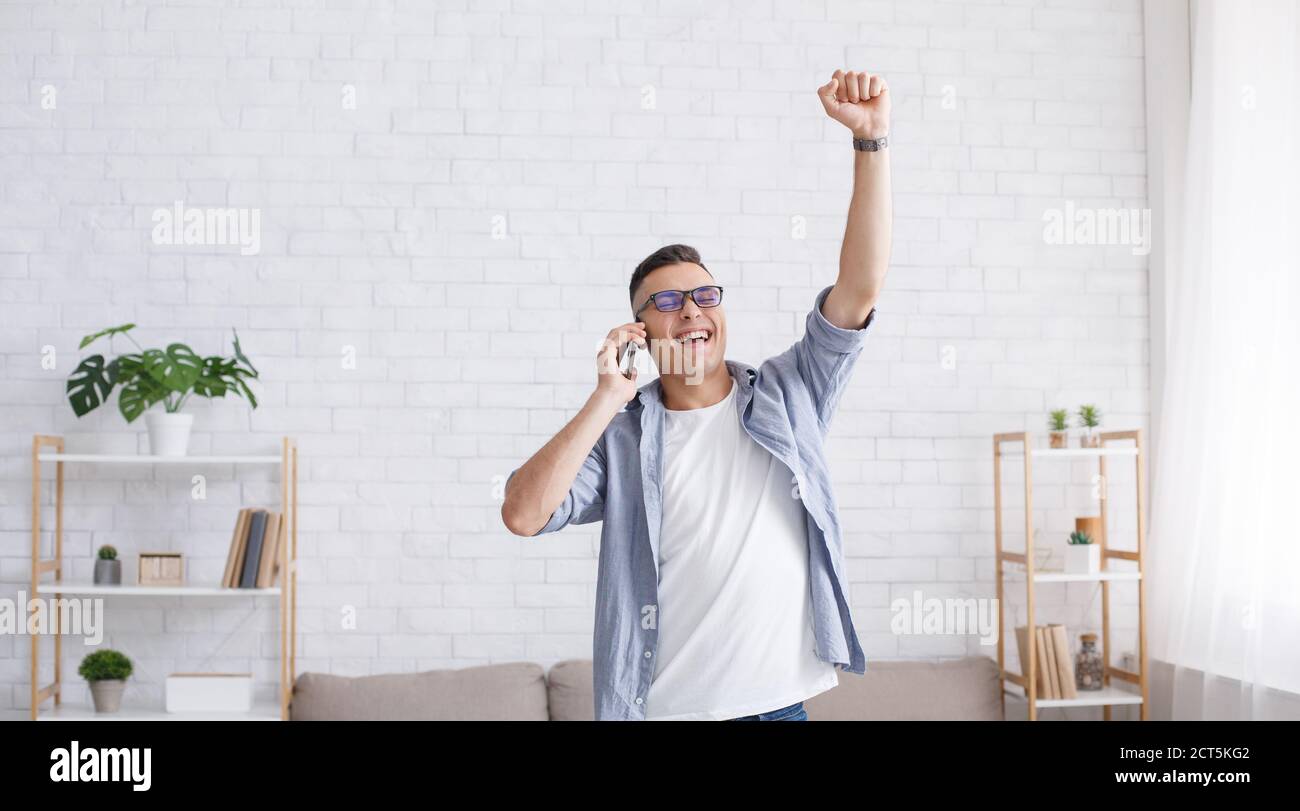 Great mood at home during remote work. Happy millennial guy with glasses got good news on smartphone call and expresses emotions of happiness Stock Photo