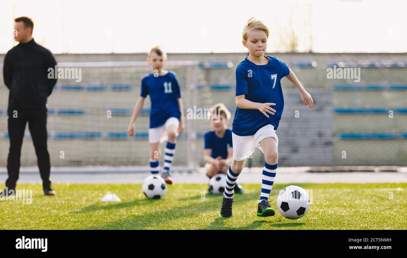 Group of School Kids with Young Coach Kicking Soccer Balls on Grass Sports Pitch. Junior Footballers in Blue Shirts on Training. Horizontal Image of S Stock Photo