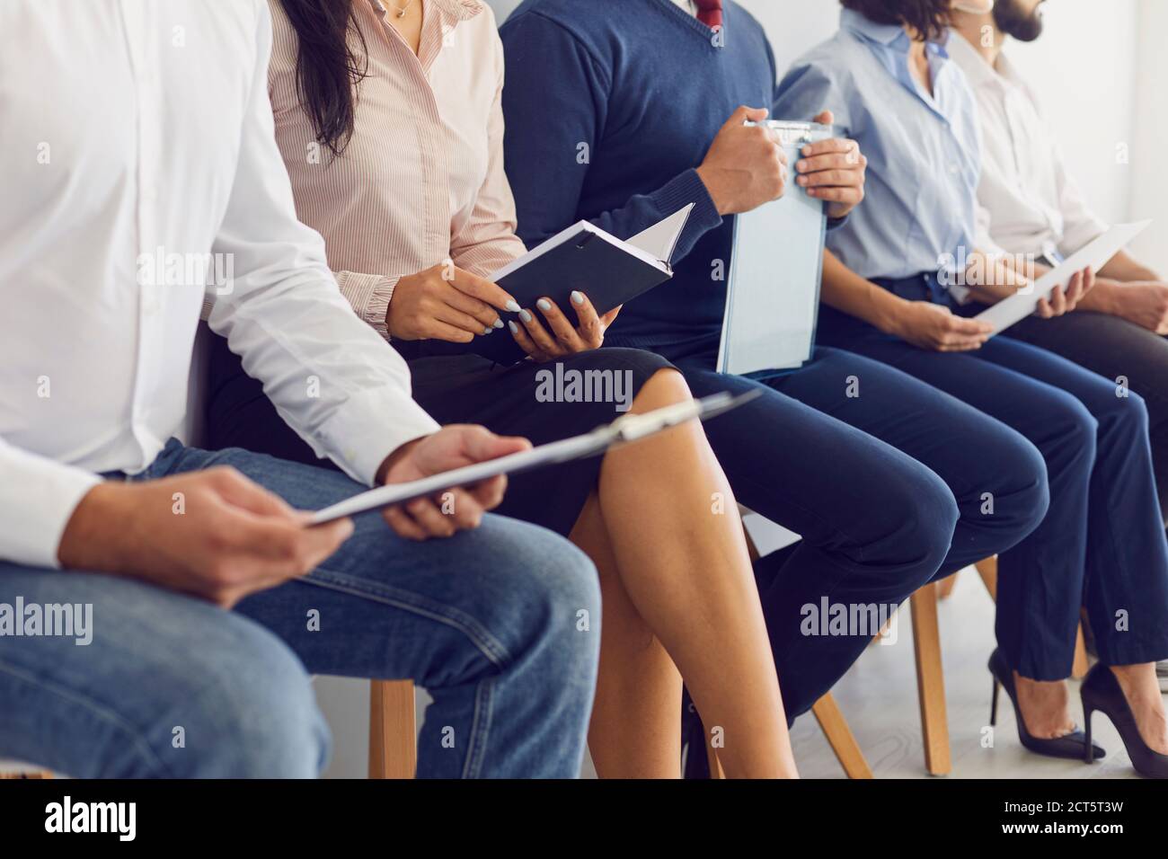 Cropped image of people sitting on chairs in a queue and holding paper in their hands. Stock Photo