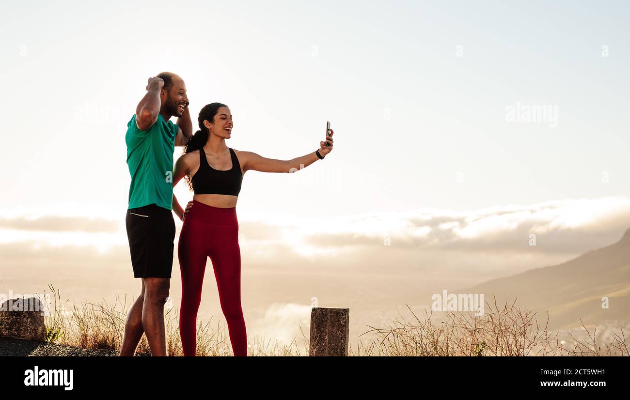 fitness couple standing outdoors taking a selfie after workout. Couple having fun relaxing after a run taking a selfie using mobile phone. Stock Photo