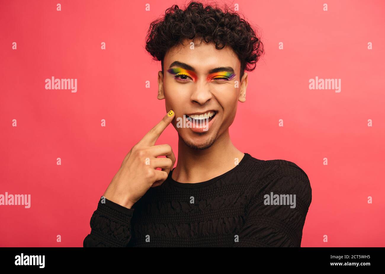 gay man wearing multicolored shadows on the eyelids winking at camera happy transgender male winking an eye against red background stock photo alamy