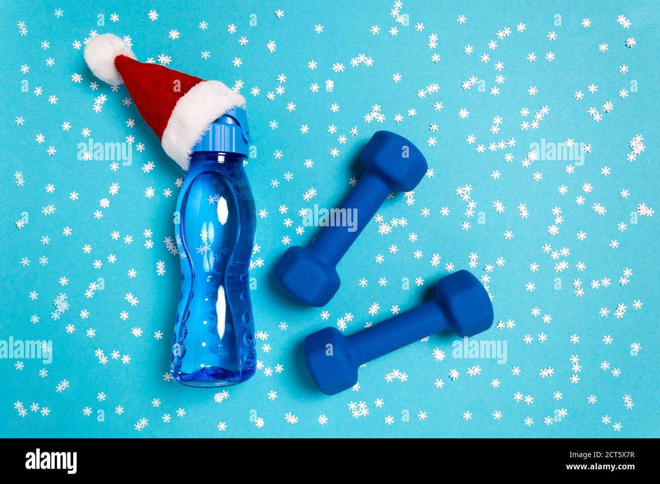 Christmas sports flat lay with dumbbells and water bottle in red Santa's hat on blue background. Christmas and new year holiday concept for fitness, w Stock Photo