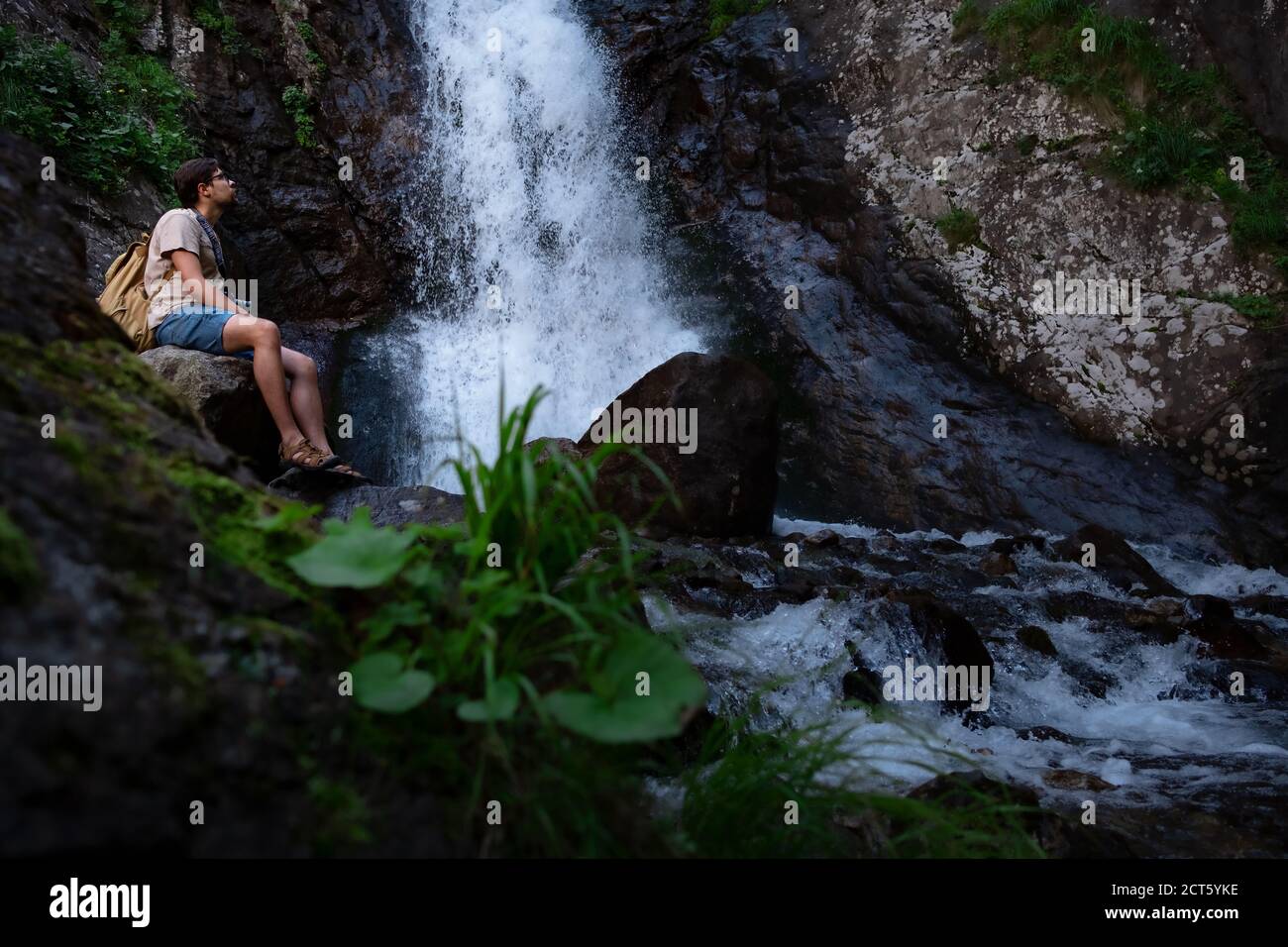 Hiker waalkinh with backpack looking at waterfall in park in beautiful nature landscape. Portrait of male adult back standing outdoor. Waterfall Shumk Stock Photo