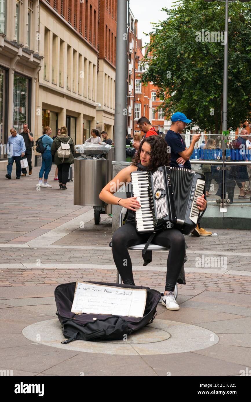 London West End Knightsbridge street scene pretty young New Zealand girl plays accordion for money busking busker Harrods fund school travel seated Stock Photo