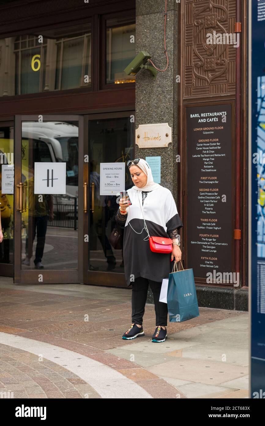 London West End Knightsbridge street scene Harrods entrance door muslim woman mobile cell phone sunglasses hijab abaya bag shopping pavement sidewalk Stock Photo