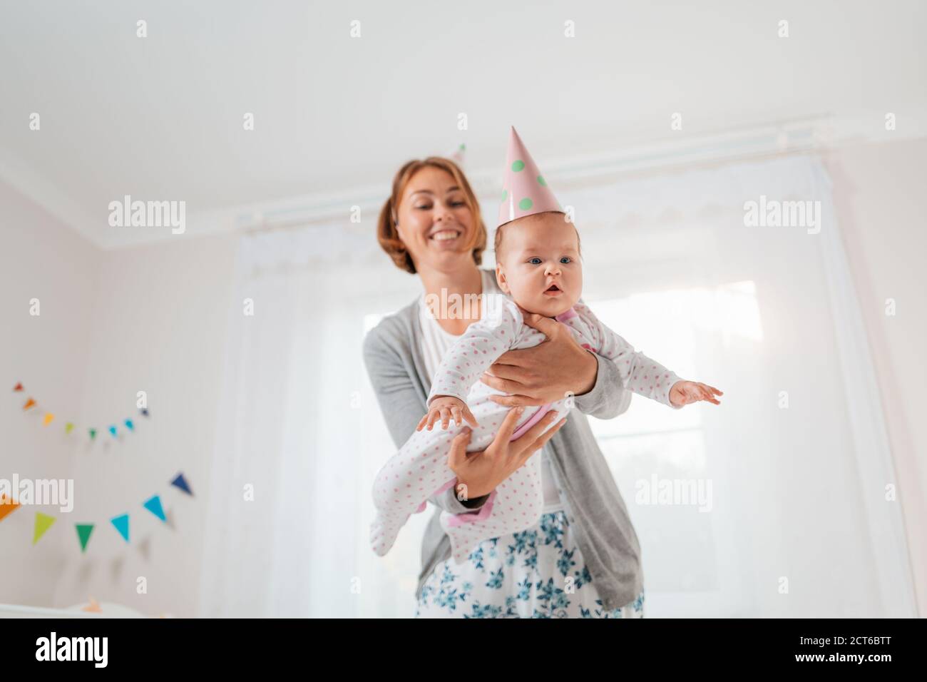 A smiling mother holds a cute baby in her arms, playing with it, imitating the flight of an airplane. Holiday hats on their heads. Concept of motherho Stock Photo