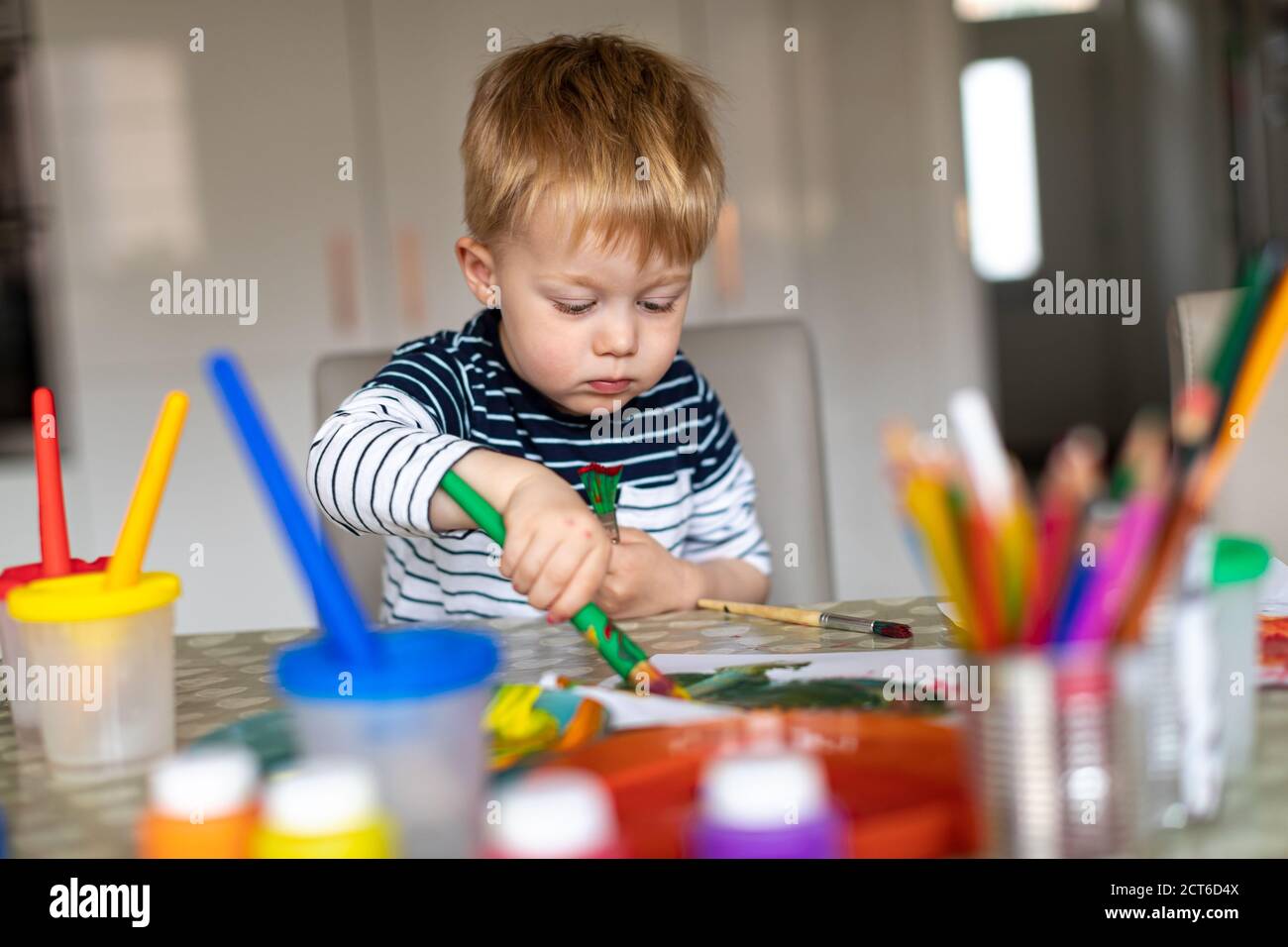 Three year old boy busy painting at home, with paint pots and brushes