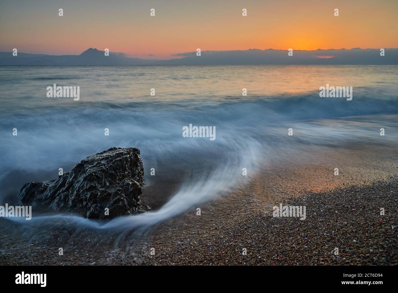 A rock stands on the pebble beach and is hit by the sea wave at sunset with very beautiful colors of the sea sky of the wave and the pebbles. The wave Stock Photo