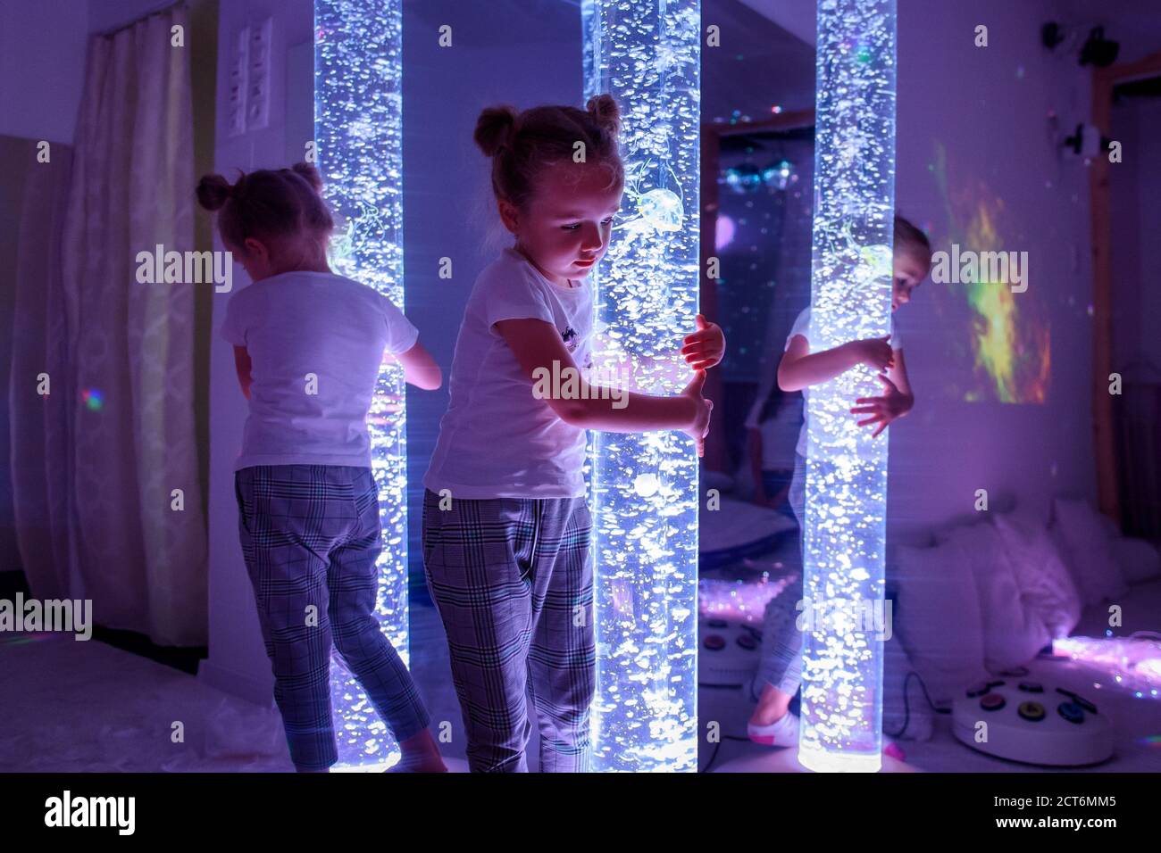 Child in therapy sensory stimulating room, snoezelen. Autistic child interacting with colored lights bubble tube lamp during therapy session. Stock Photo