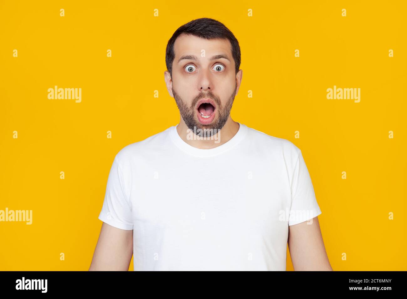 Young man with beard in white t-shirt with open mouth, afraid and shocked by unexpected expression, fear and excited face, stands on isolated yellow background. Stock Photo