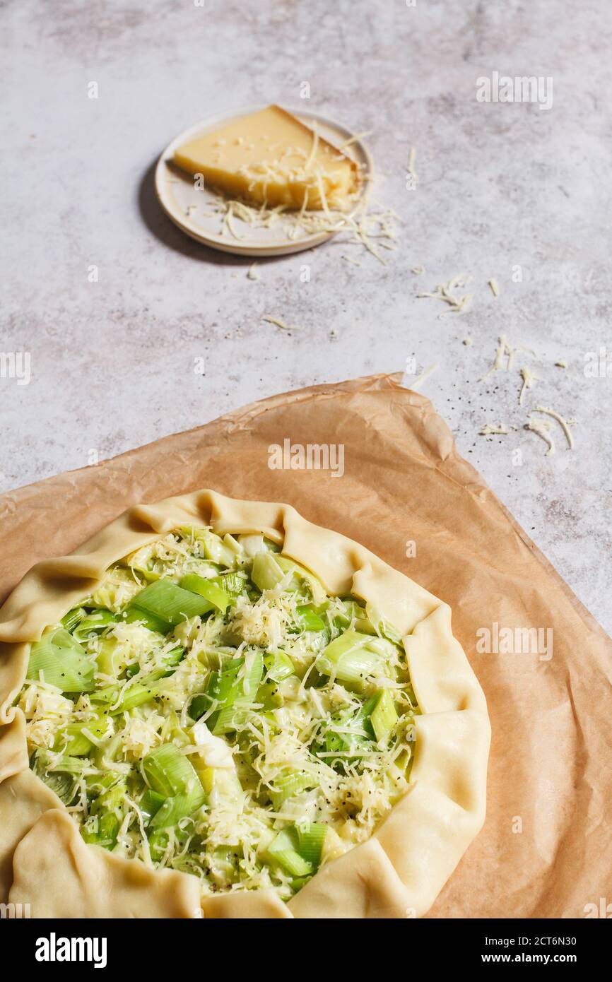 Making a leek and cheese galette. Raw savoury pastry being prepared on a piece of baking paper with a small plate of grated cheese in the background. Stock Photo