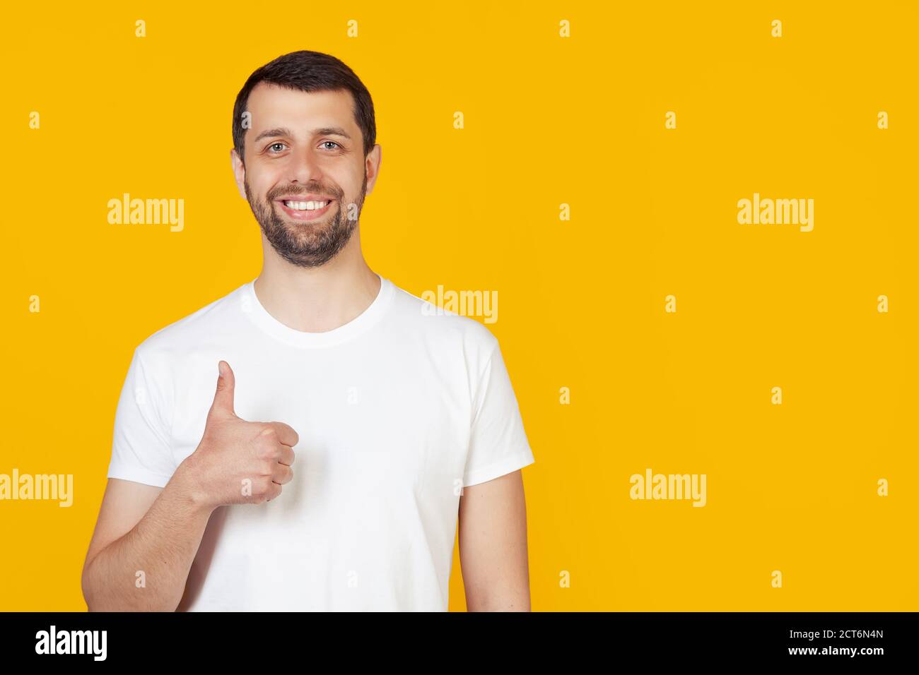 Young man with a beard in a white t-shirt making a happy hand gesture. Confirming facial expression showing success while looking at the camera. Stands on isolated yellow background Stock Photo