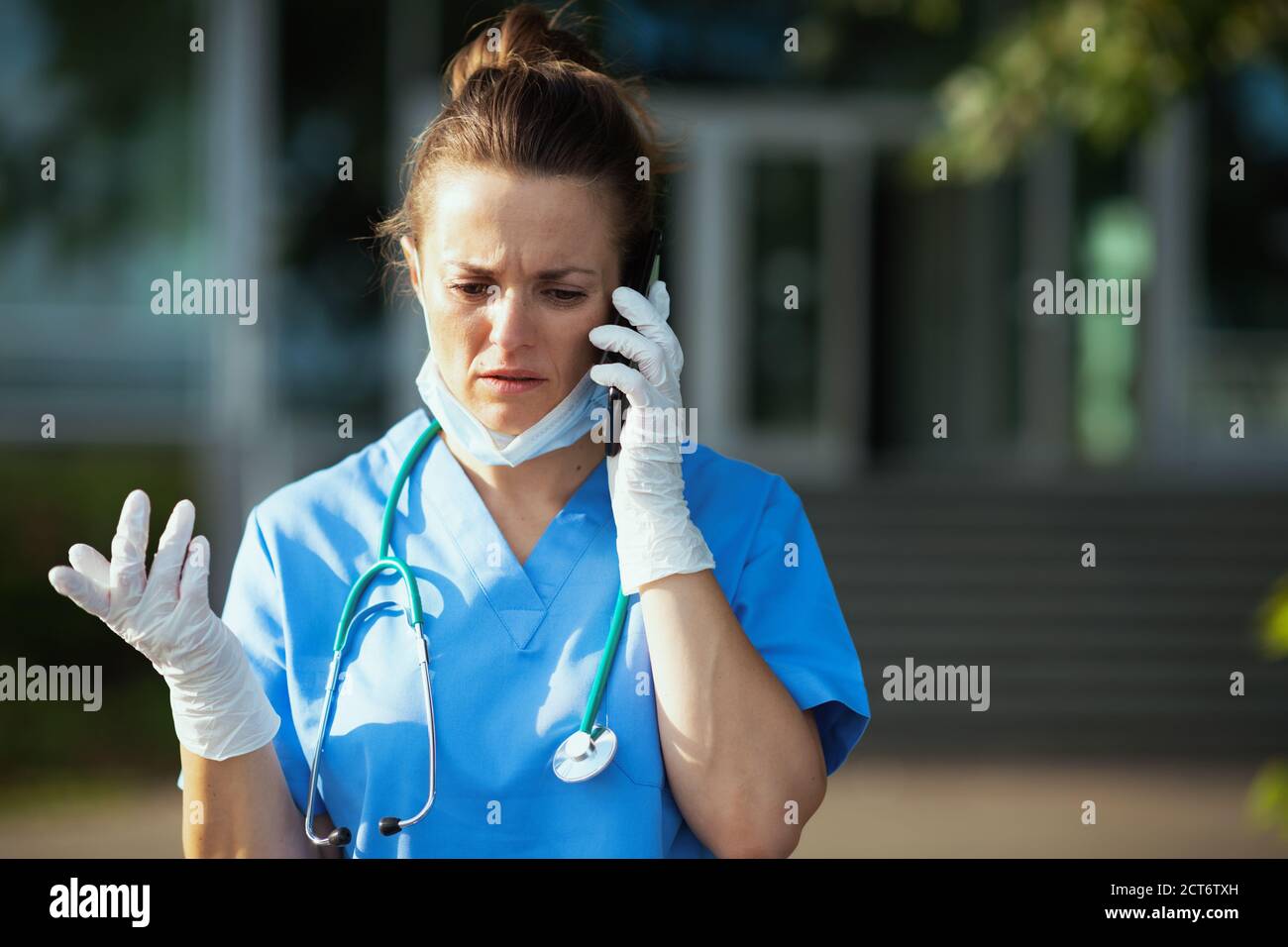 coronavirus pandemic. unhappy modern physician woman in uniform with stethoscope and medical mask using a smartphone outdoors near clinic. Stock Photo