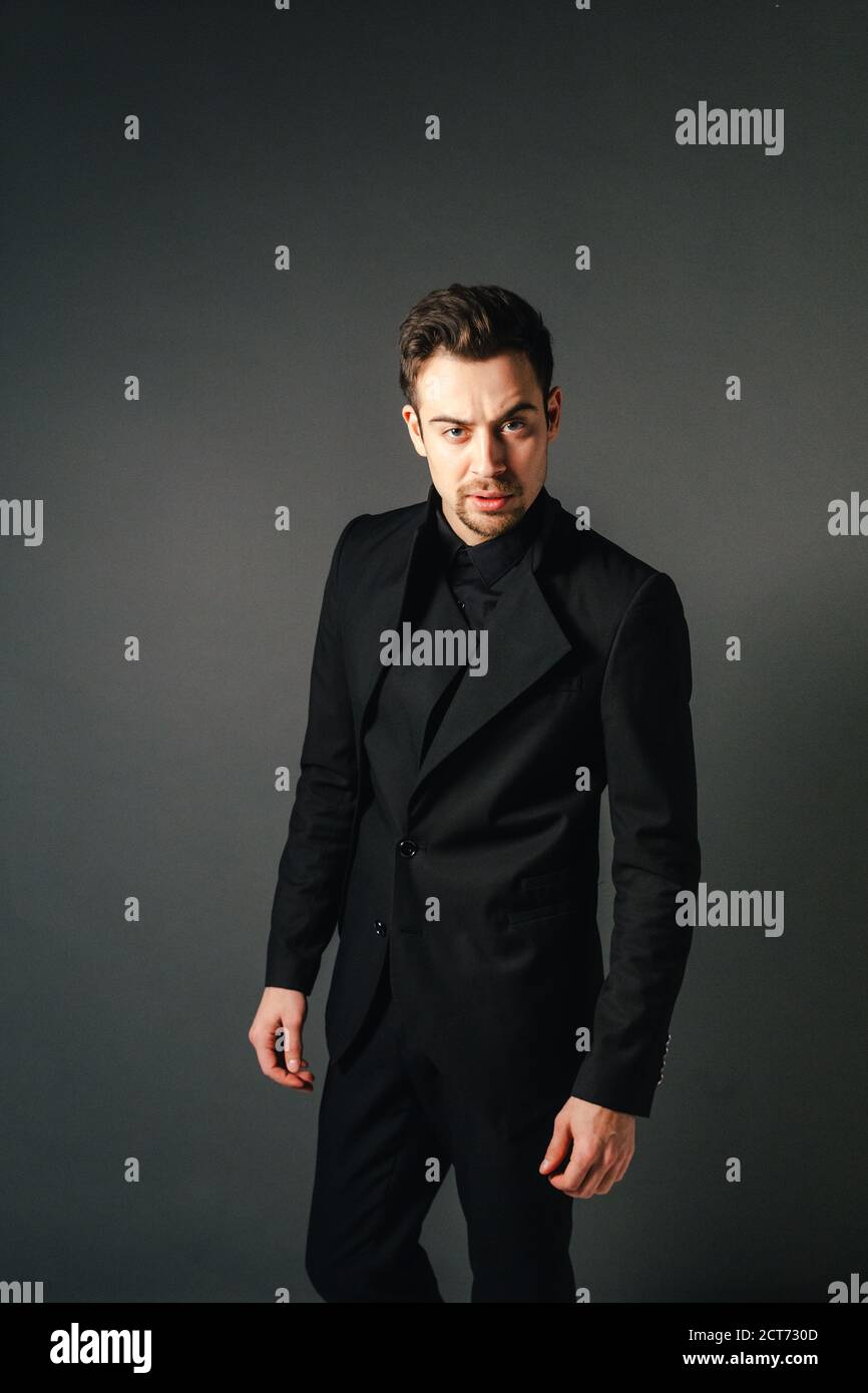 Young handsome man in a black suit, standing and seriously looking at the camera, against plain studio background Stock Photo