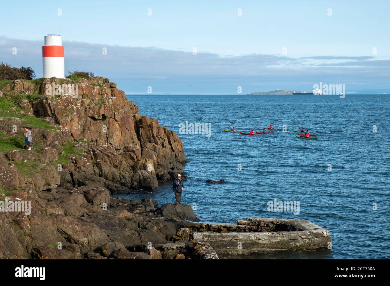Sea Kayakers pass as people fish from a rocky outcrop on the Fife coastline near Aberdour, Scotland. Stock Photo