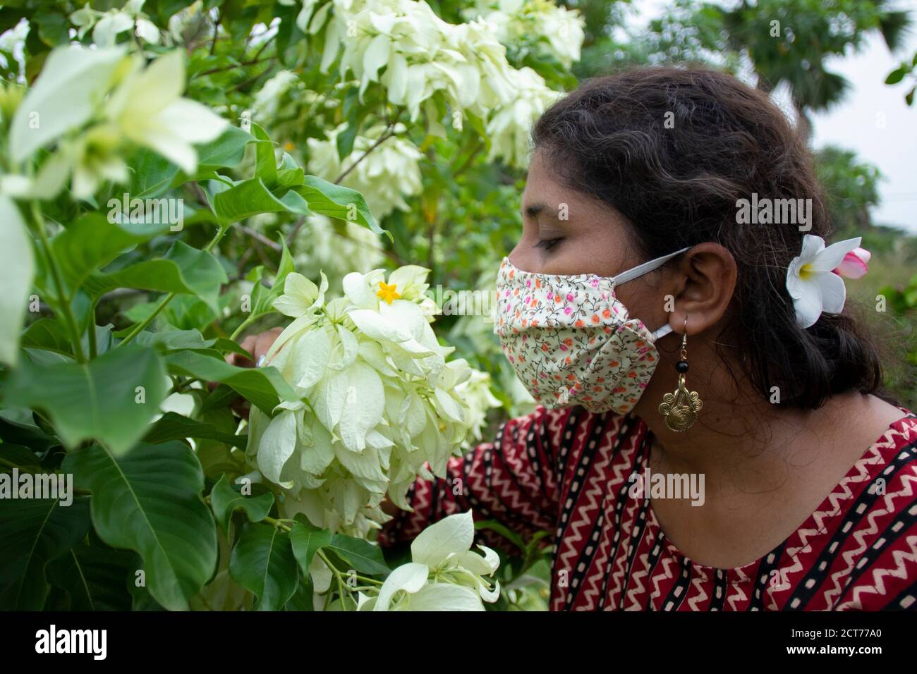 Beautiful lady with floral mask, earrings and designer top smells white Mussaenda flowers. Protection from corona in a stylish manner. Stock Photo