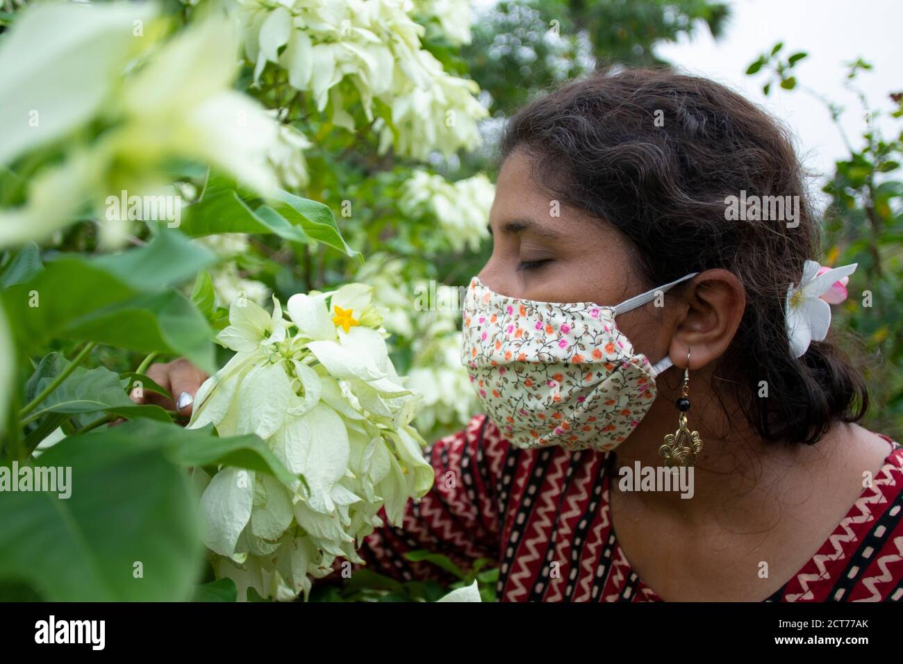 Beautiful lady with floral mask, earrings and designer top smells white Mussaenda flowers. Protection from corona in a stylish manner. Stock Photo