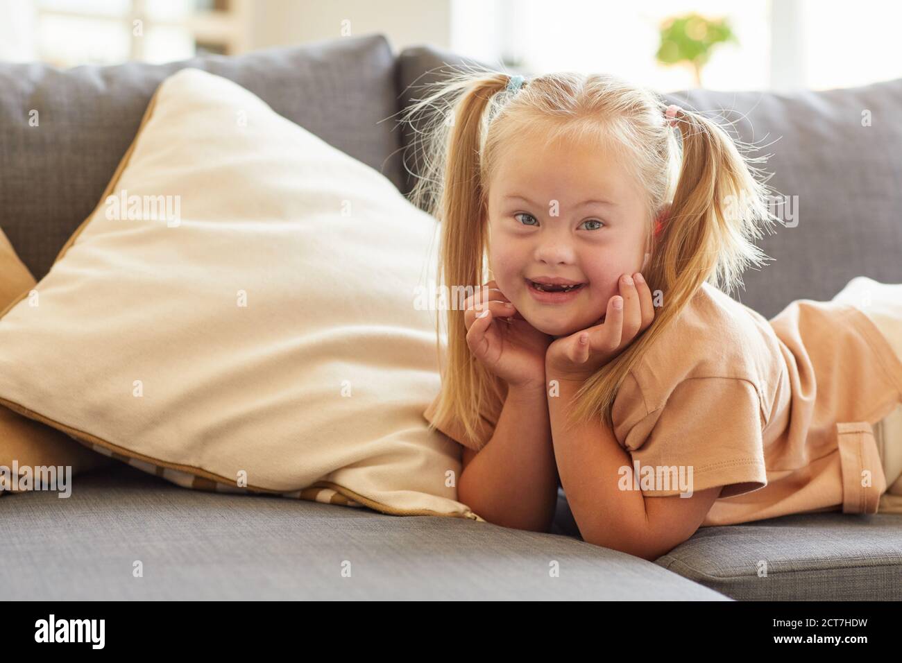 Portrait of cute girl with down syndrome smiling happily at camera while lying on sofa at home, copy space Stock Photo