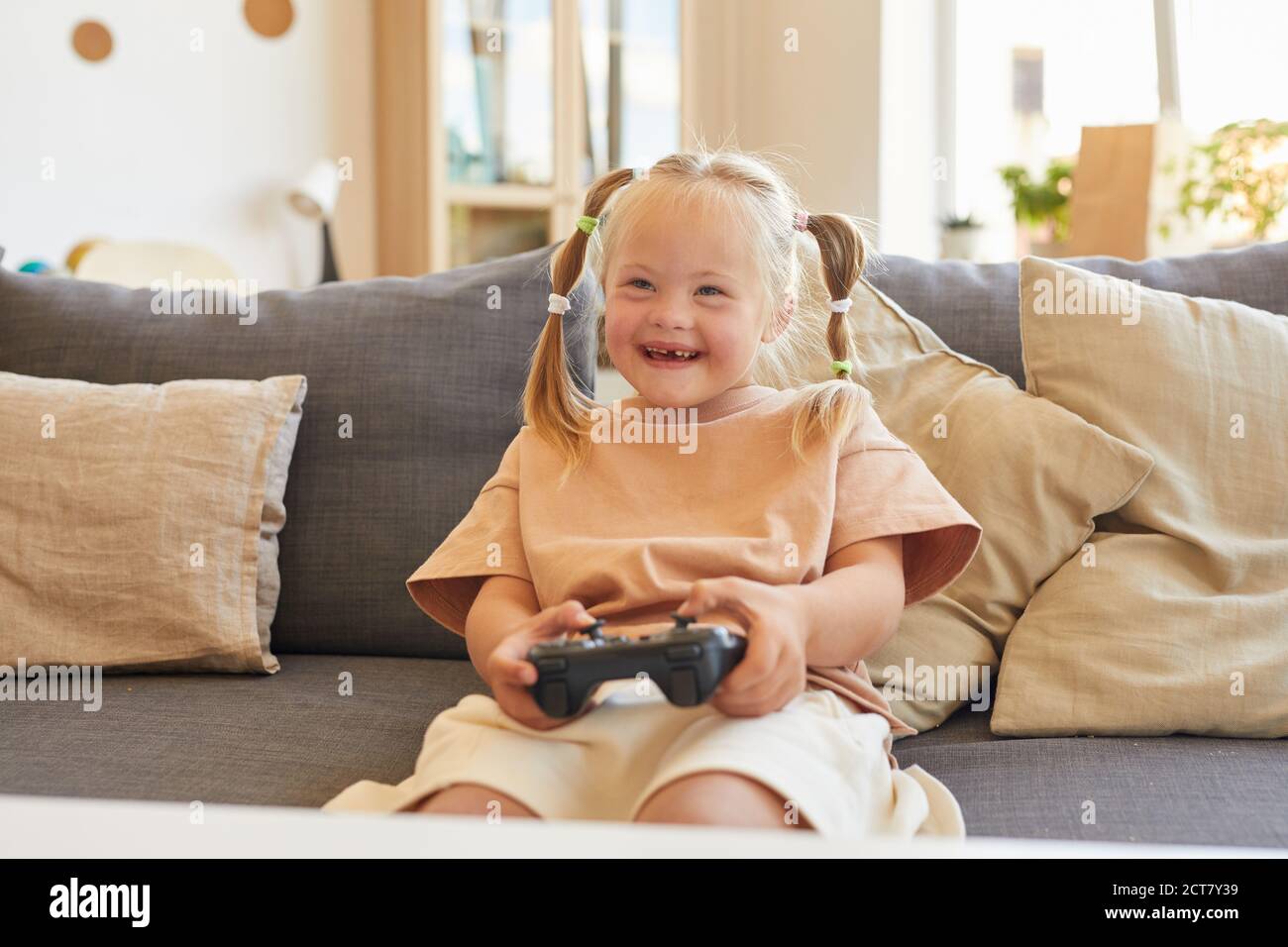 Portrait of cute girl with down syndrome playing video games and laughing happily while sitting on couch in living room, copy space Stock Photo