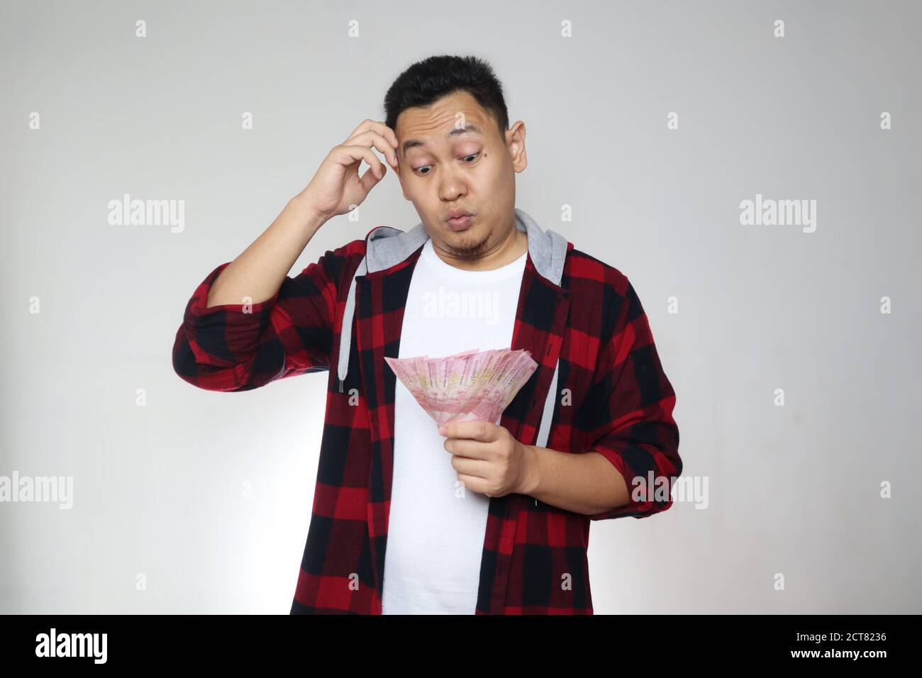 Confused stressed Asian man holding Indonesian rupiah cash in hand, thinking wondering how to spend. More money more problems  or not enough money con Stock Photo