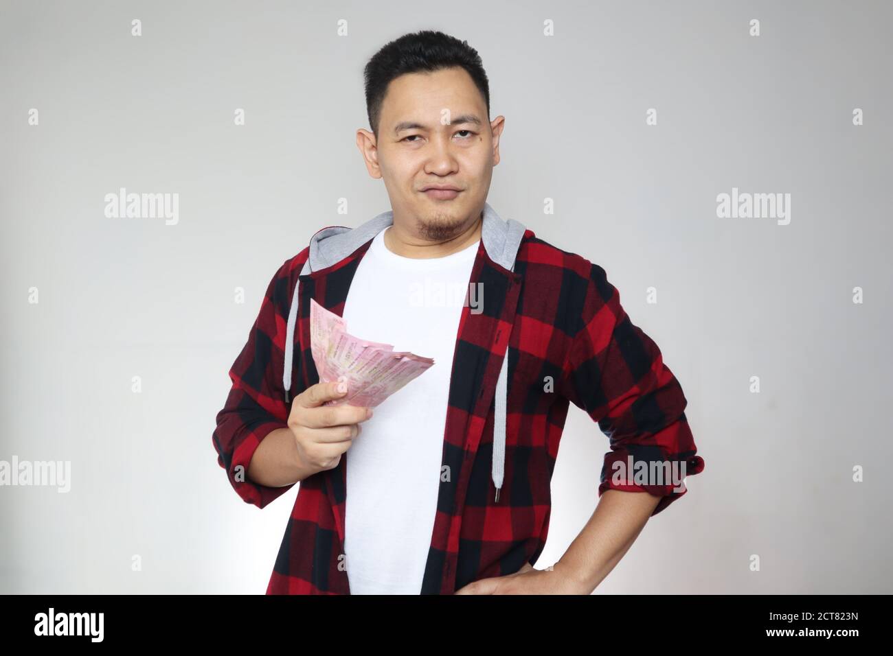 Portrait of happy young Indonesian man holding rupiah money, smiling laughing winning gesture, over white background Stock Photo