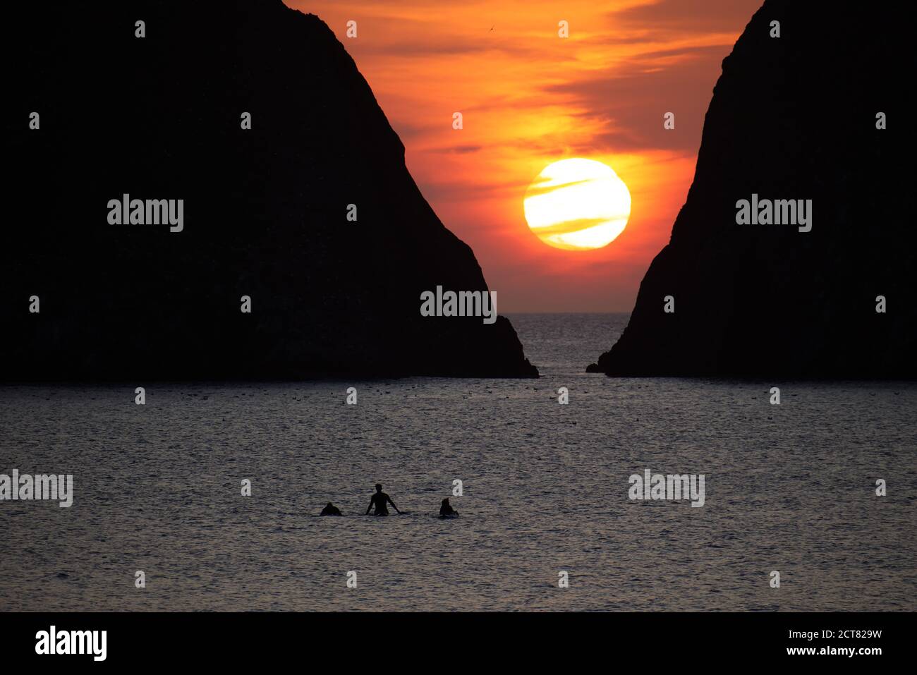 Three silhouetted unidentifiable surfers return from the sea in front of Gull Rocks at Holywell Bay in Cornwall as the sun sets behind the clouds. Stock Photo