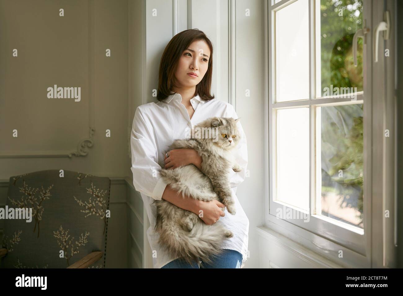beautiful young asian woman standing by window at home holding a cat in arms looking depressed and sad Stock Photo