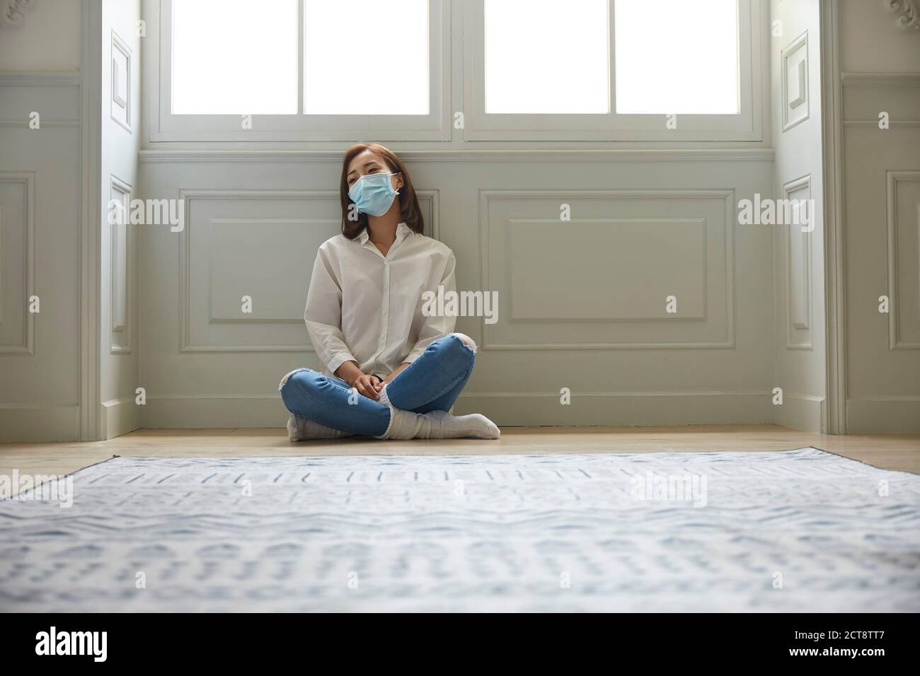young asian woman in quarantine at home wearing facial mask sitting on floor legs crossed looking sad and depressed Stock Photo