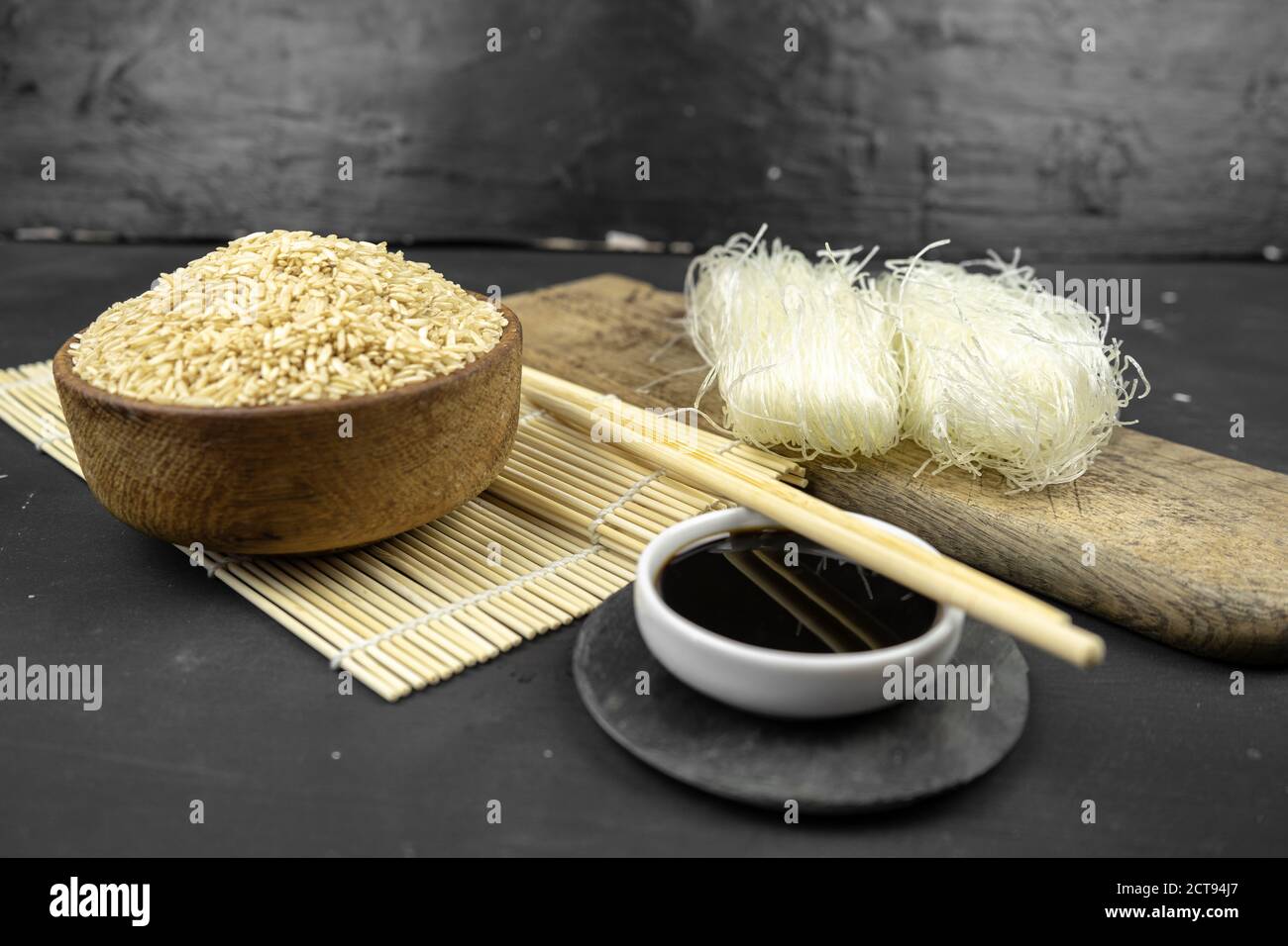 brown rice in a wooden bowl, asian kitchen background with bamboo mat, chopsticks, soy sauce, pickled ginger and glass rice noodles on black backgroun Stock Photo