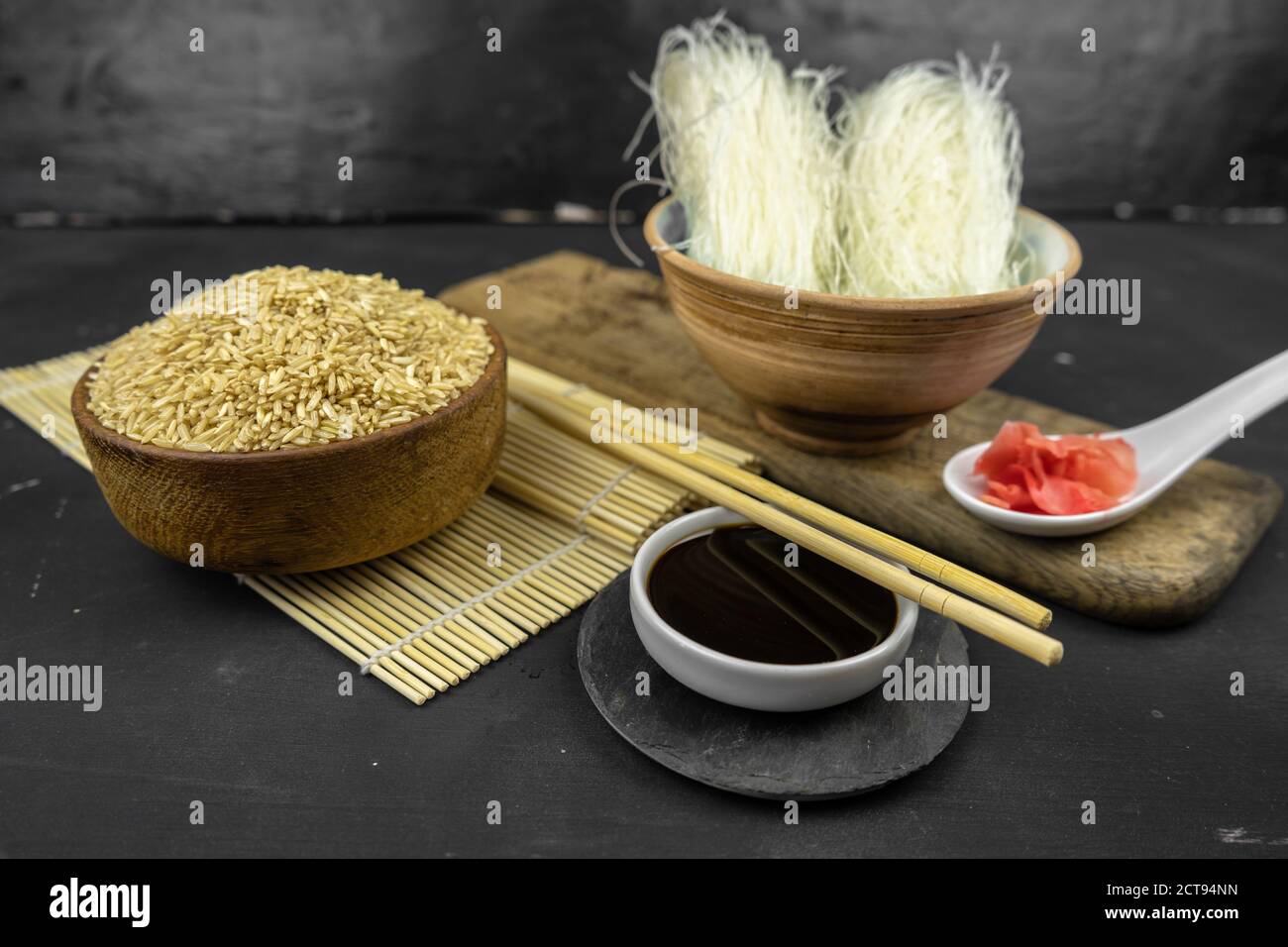 brown rice in a wooden bowl, asian kitchen background with bamboo mat, chopsticks, soy sauce and glass rice noodles on black background Stock Photo