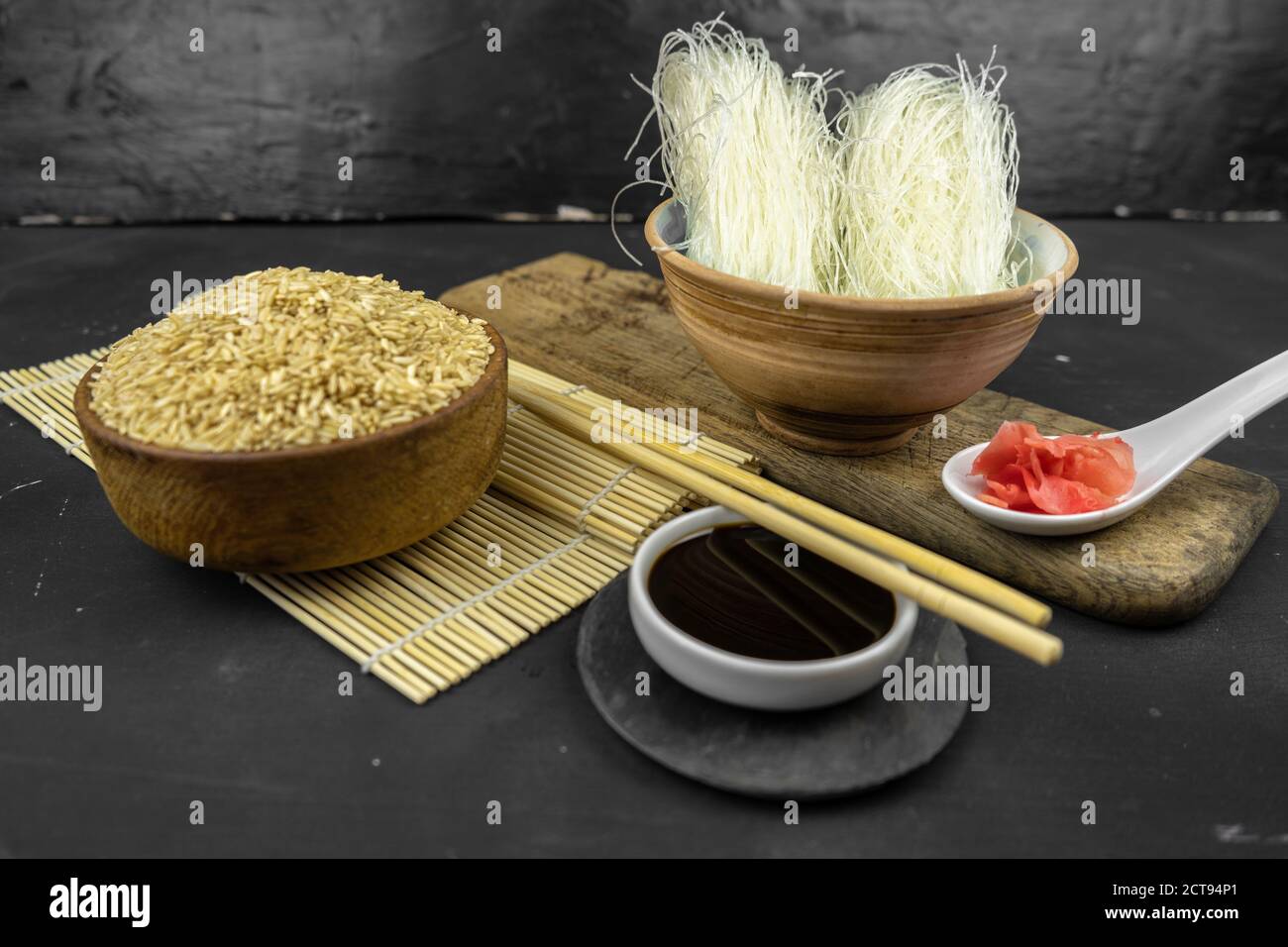 brown rice in a wooden bowl, asian kitchen background with bamboo mat, chopsticks, soy sauce and glass rice noodles on black background Stock Photo
