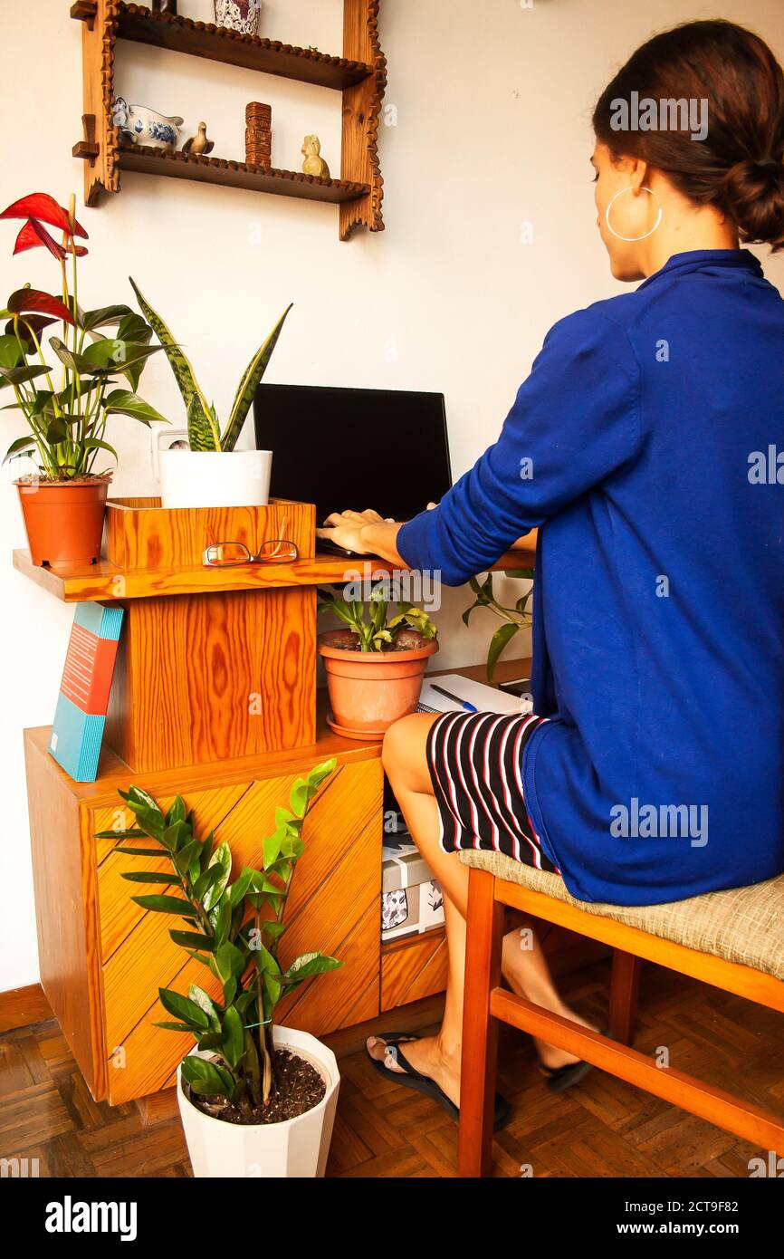 Young woman teleworking from home sitting in her desk decor with plants. Writing in a computer. Long full shot. Stock Photo