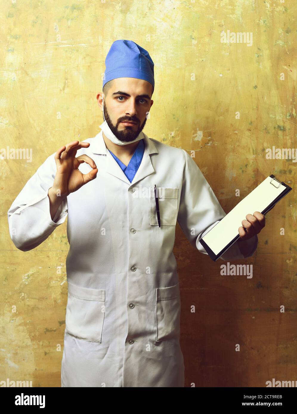 Doctor with dark beard and assured face expression shows ok sign and holds clip folder in hand on beige background. Concept of medicine, professionalism and recovery Stock Photo