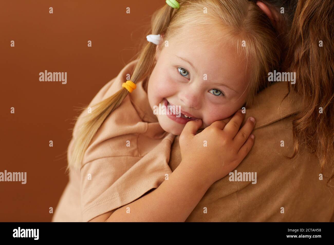 Close up of mother holding happy daughter with down syndrome while standing against plain brown background in studio Stock Photo