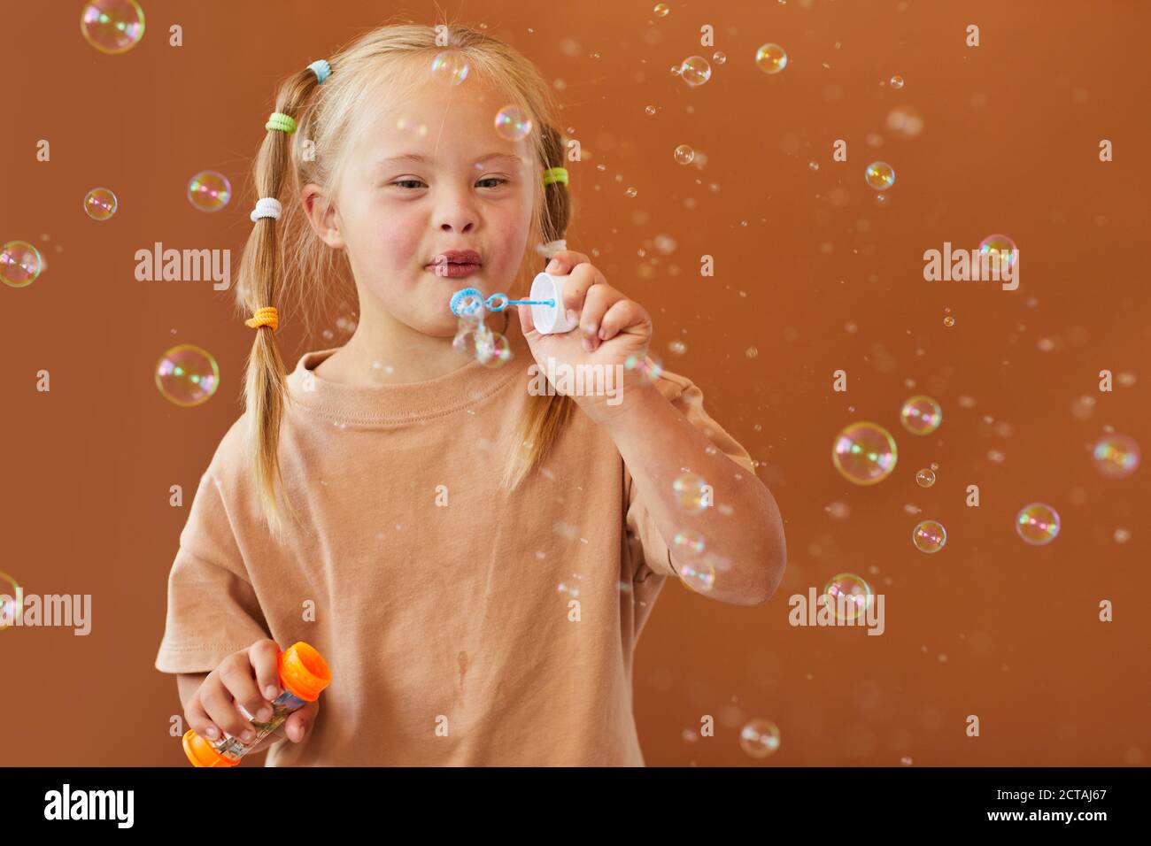 Waist up portrait of cute girl with down syndrome blowing bubbles while posing against brown background in studio, copy space Stock Photo