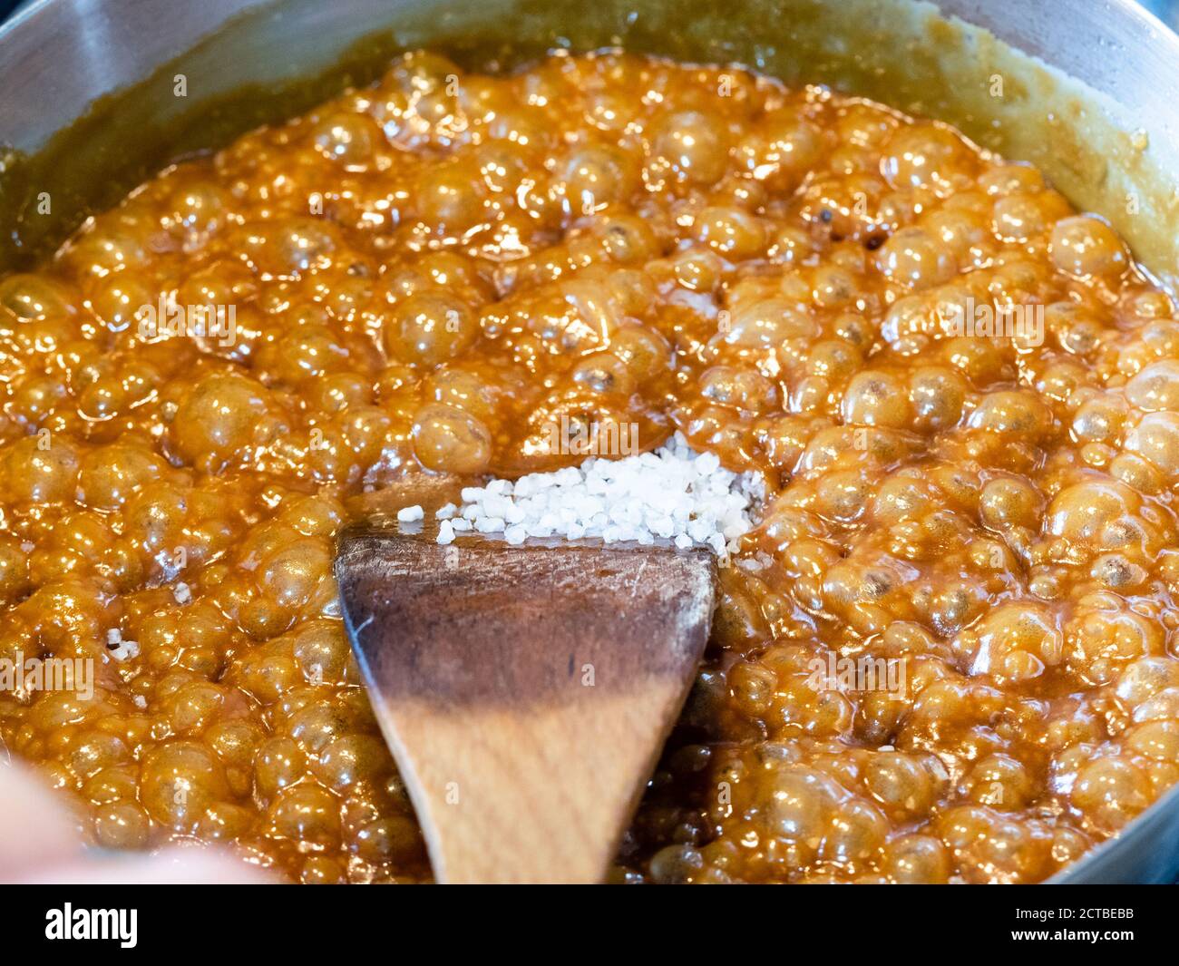 cooking salted butter caramel sauce at home kitchen - adding coarse sea salt in boiling melted sugar, butter and cream in steel pan close up Stock Photo