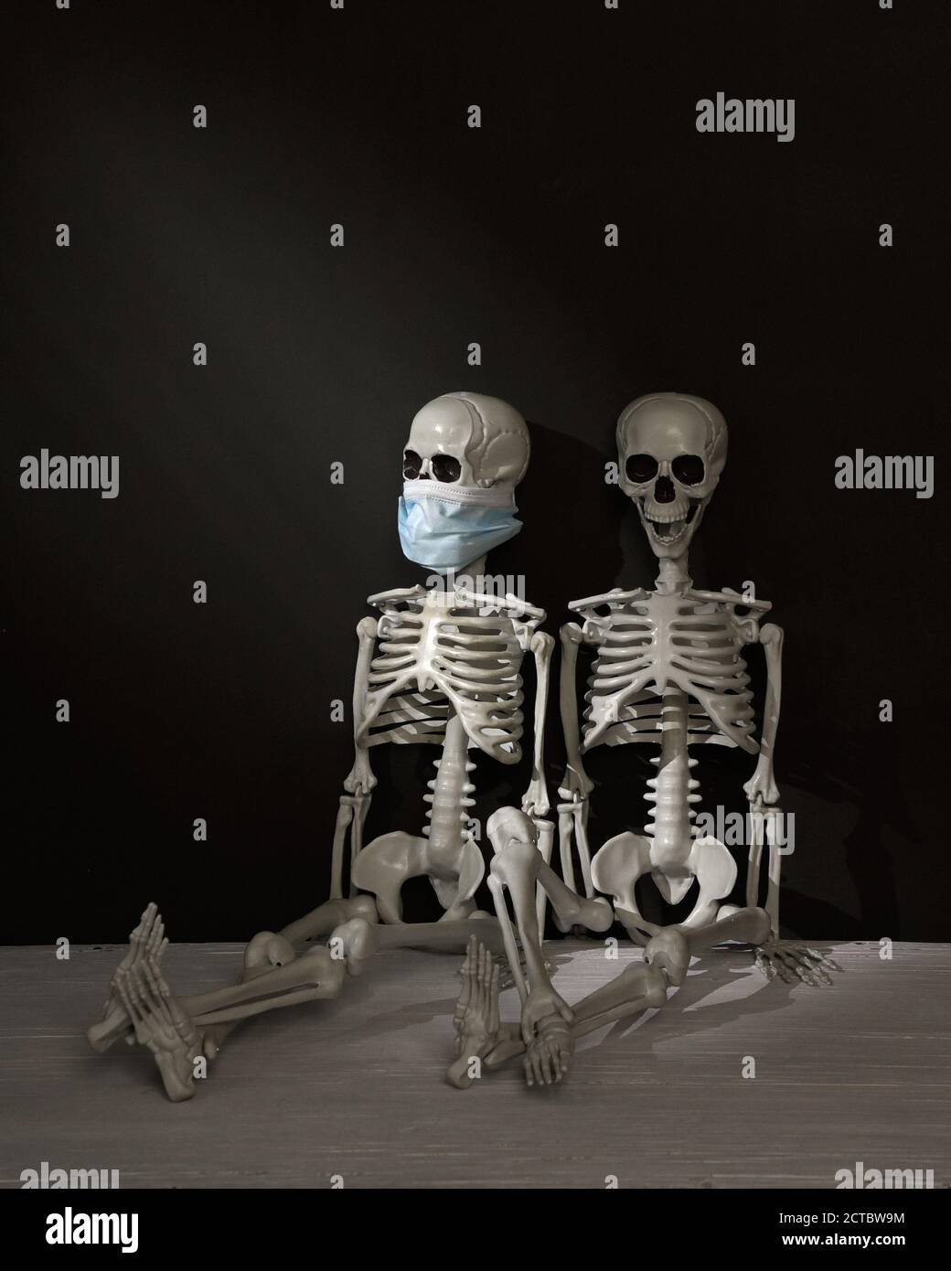 Two Skeletons are sitting in a dark room. One is wearing a mask and the other isn't for a dark humor anti mask concept during the covid 19 pandemic. Stock Photo