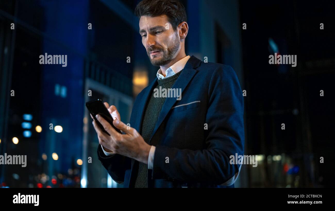 Tall Caucasian Businessman in a Suit is Using a Smartphone on Dark Street in the Evening. He's Browsing While Looks Confident and Successful Stock Photo