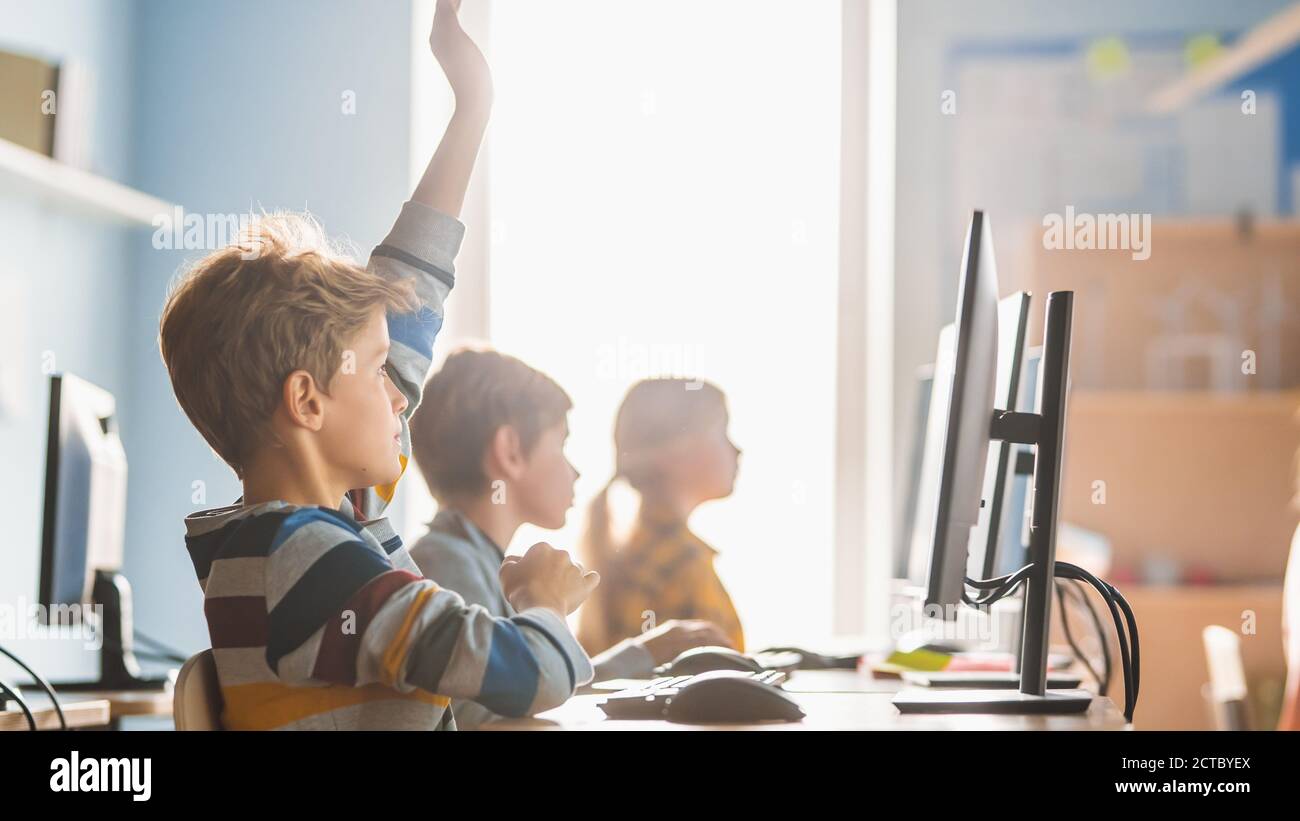 Elementary School Science Classroom: Boy Raises Hand with Question. Teacher Educates Smart Little Schoolchildren who Work on Personal Computers, Learn Stock Photo