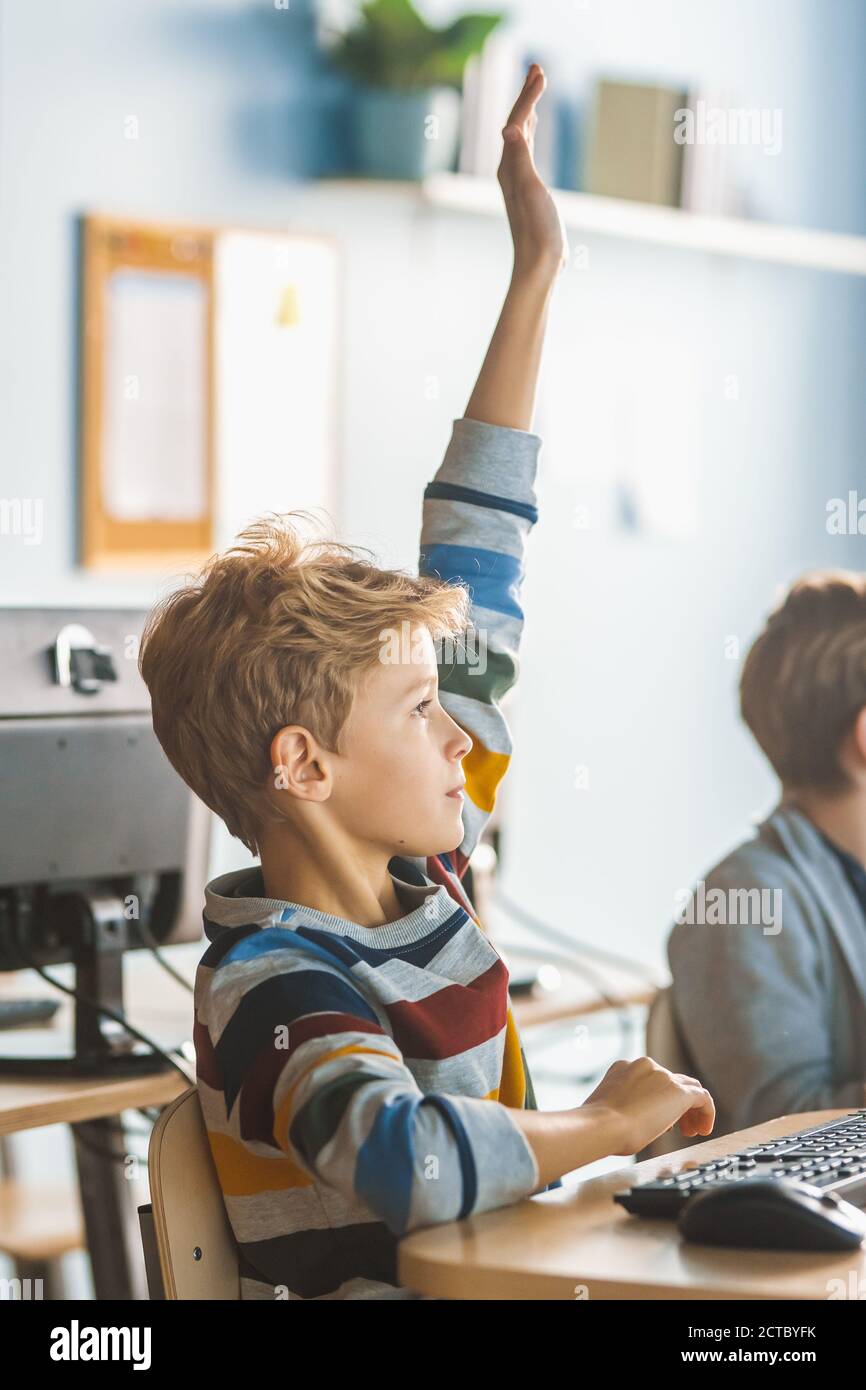 School Science Classroom: Boy Raises Hand with Question. Teacher Educates Smart Little Schoolchildren who Work on Personal Computers, Learn Stock Photo
