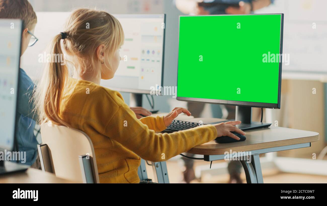 Elementary School Computer Science Classroom: Cute Little Girl Uses Green Mock-up Screen Computer while Learning Coding and Programming Stock Photo